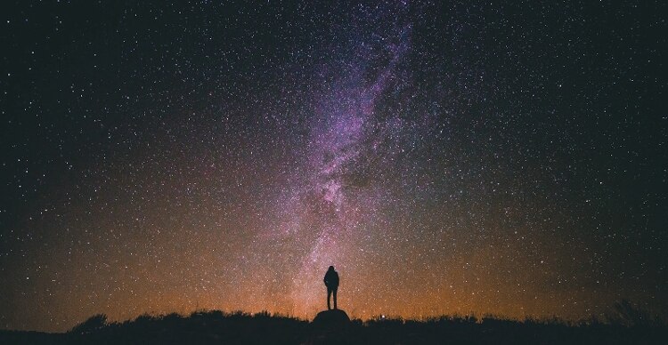black silhouette of a person standing on a rock under a dark starry sky with large cluster of stars illuminated in shades of purple and orange in middle