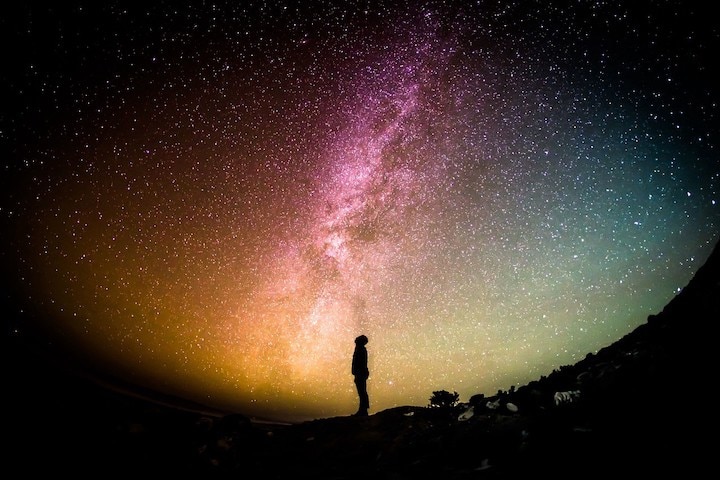 black silhouette looking up at galaxy sky with white stars and tones of pink, green, blue and yellow