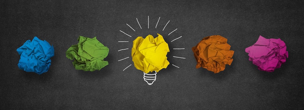 blackboard with 5 crumbled up pieces of paper - blue, green, yellow, orange, and pink. yellow one in center has white chalk lines around it to create a lightbulb