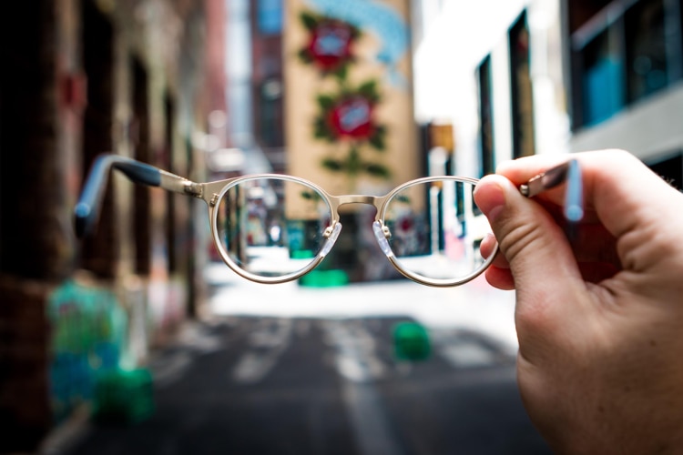 pair of glasses held up to blurry background of what appears to be a street with colored building walls
