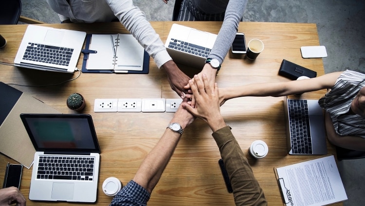group of men and women of different races sitting around light brown wooden desk with open silver laptops w/ black keyboards. there is a white notebook, black wallet, and black cups of coffee around the table and a white outlet bank in the middle of the table. everyone in group is extending their hand on top of each other above the table, to indicate teamwork