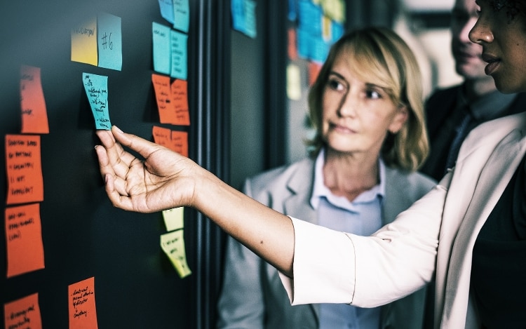 red, blue and yellow sticky notes with black text posted on a wall. a woman's arm (with light pink suit sleeve) is outstretched, and holding onto the blue sticky note. a woman with blond hair is watching in the background.