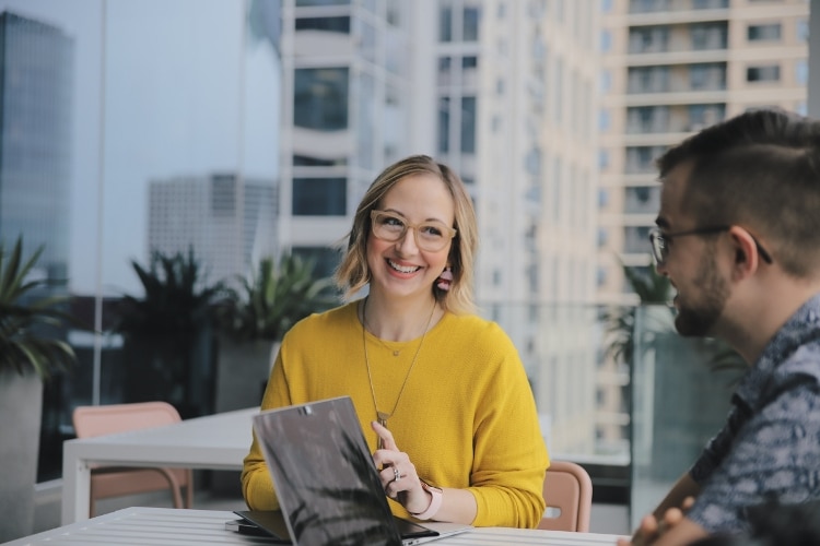 woman with short blonde hair and glasses, wearing yellow shirt, smiling at a man wearing glasses and a blue collared shirt. reader can only see man from behind/side angle.