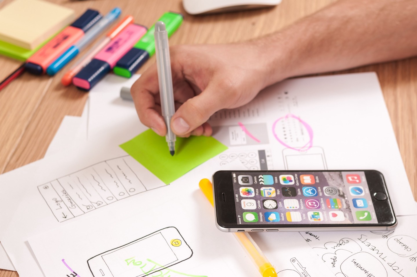 iphone resting on desk with scattered papers and blue, pink, yellow, and green highlighters. man's hand is holding grey marker and about to write on a green sticky note next to the iphone