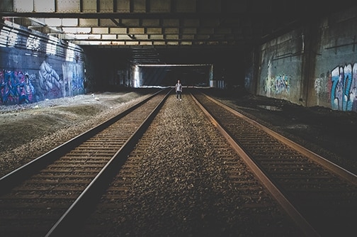 2 empty train tracks with figure of a man in between them and graffitied brick walls on each side
