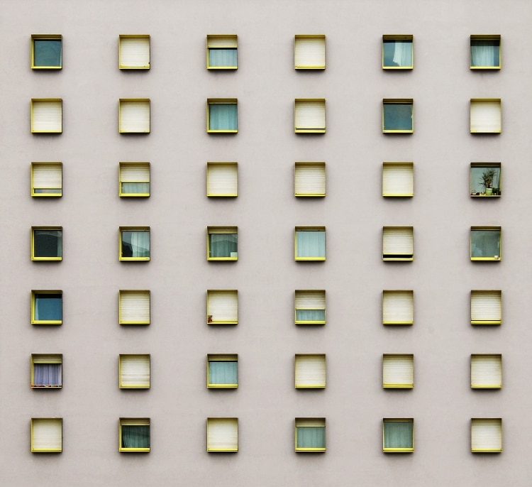 tan wall with rows of windows outlined in yellow. each window is filled with a different color - white, shades of green, or blue