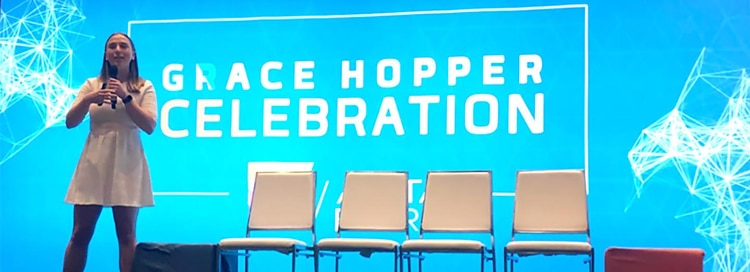 woman wearing white dress and holding microphone in front of 4 white chairs and turquoise-lit screen with white vector designs in corners and white text in middle