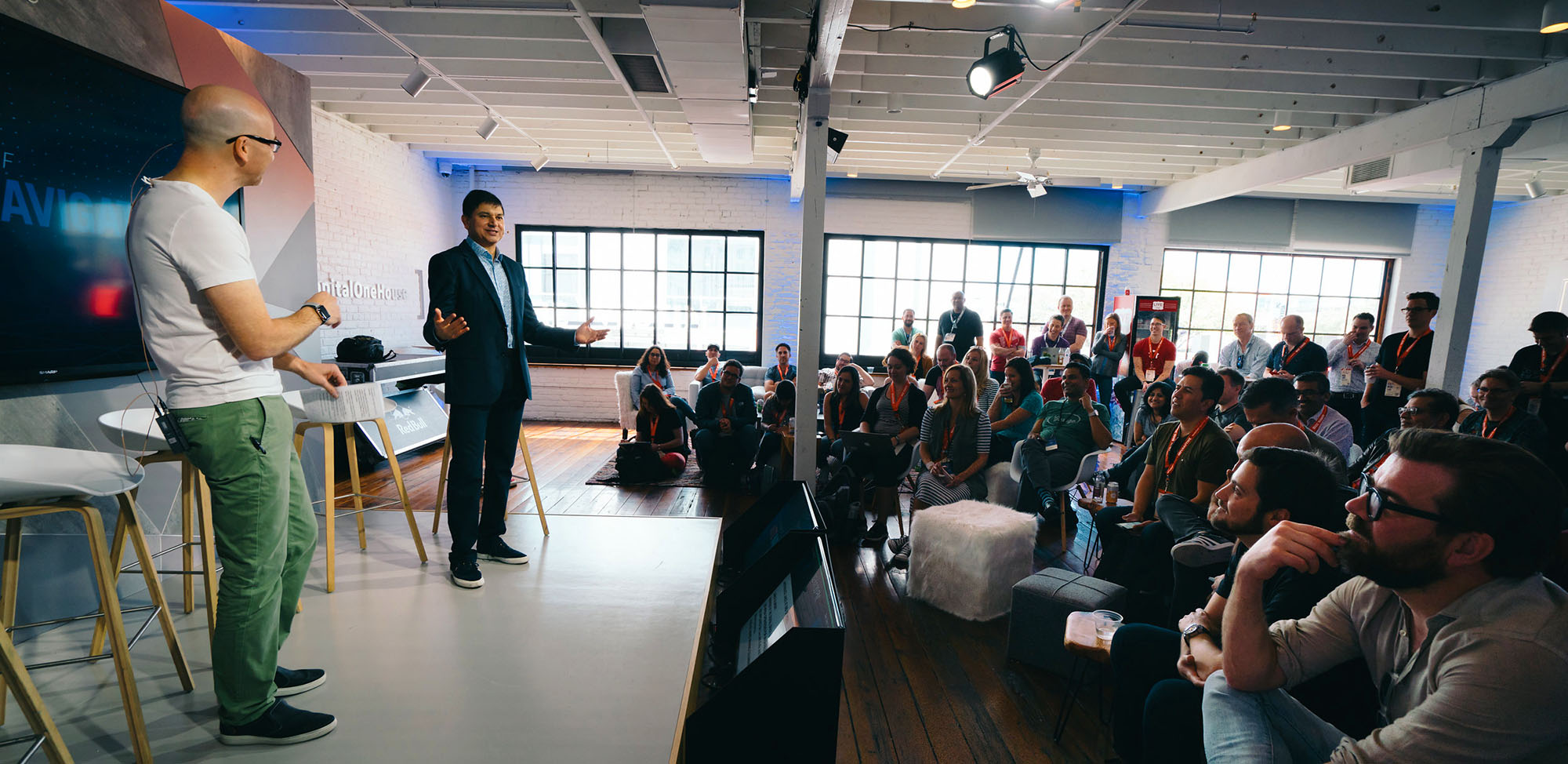 man in blue button down and navy sports jacket standing on stage and talking to a group of associates dressed in casual business wear and orange lanyards. there is another man on stage wearing a white t-shirt and green pants, directing the man lecturing.