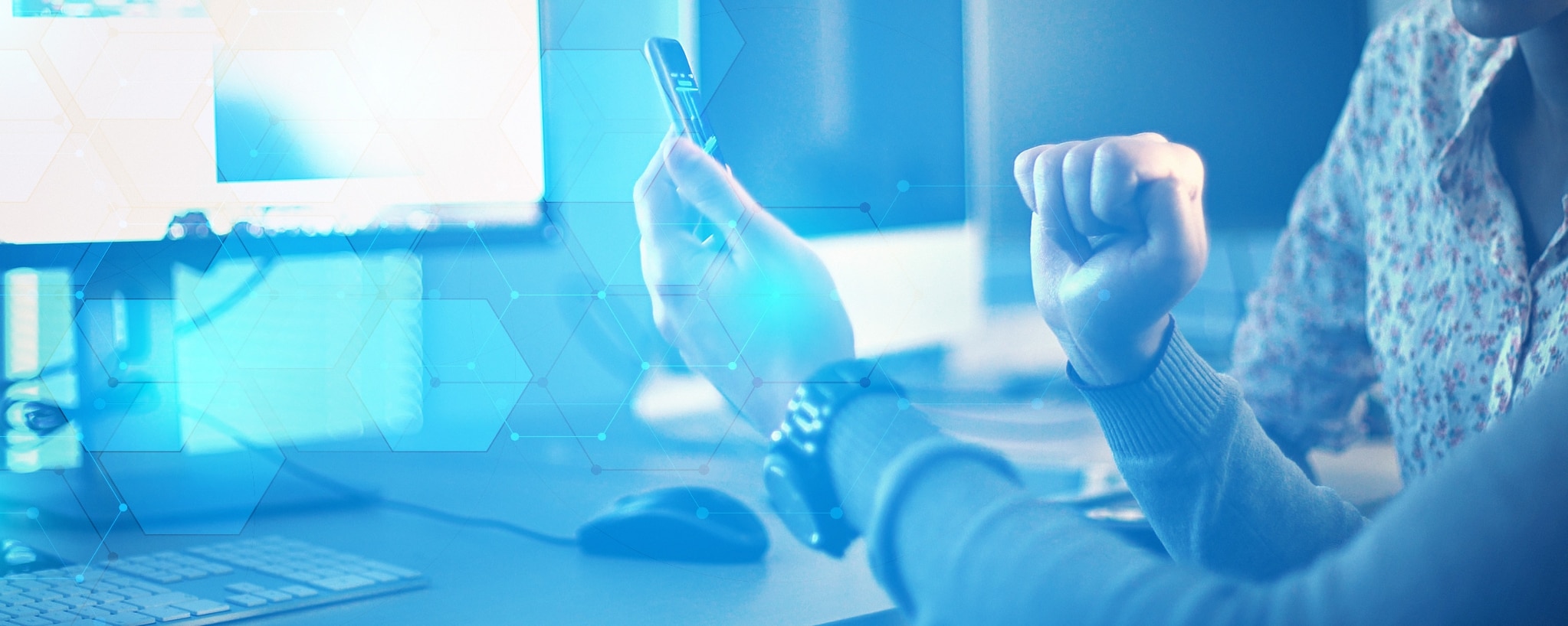 close up of man's hands holding a phone in front of a desk with a computer on it. there is a silver watch on the left wrist. there is a blue image treatment and transparent light blue and white hexagons overlaid on top of the image