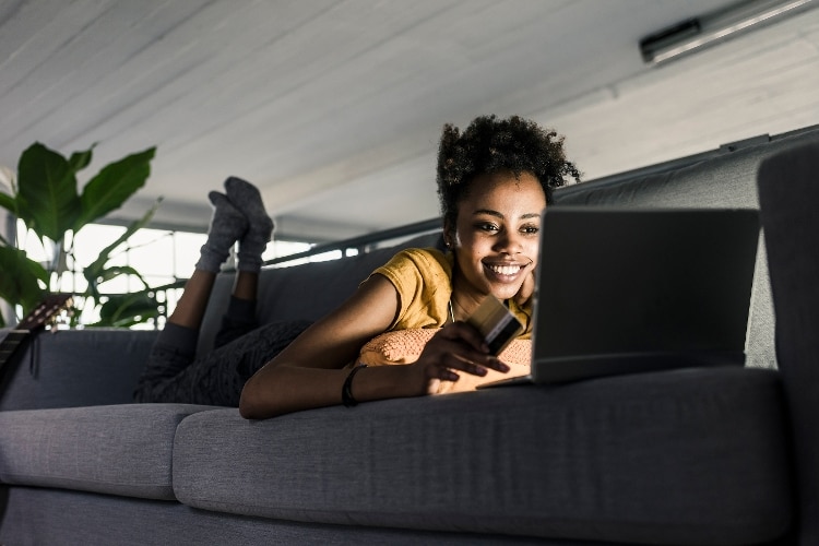 woman lying on her stomach on a grey couch, with feet in air behind her and wearing yellow shirt. she is smiling and her face is illuminated from light coming from screen of a silver laptop. she is holding a tan credit card with black stripe.