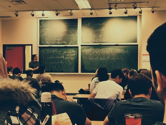classroom with man lecturing in front of 4 green chalkboards with white writing on them