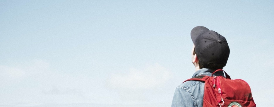 Washed out photo of a person in a black cap with a jean jacket and red backpack staring at an unseen horizon.