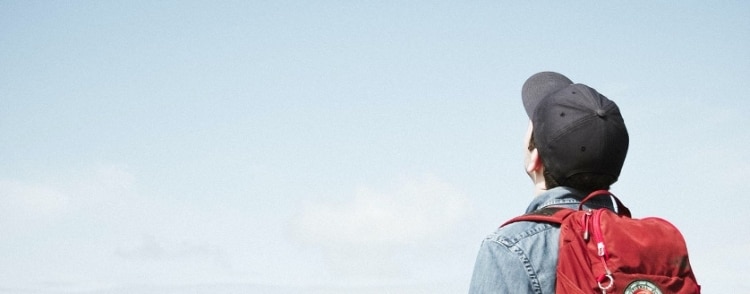 Washed out photo of a person in a black cap with a jean jacket and red backpack staring at an unseen horizon.
