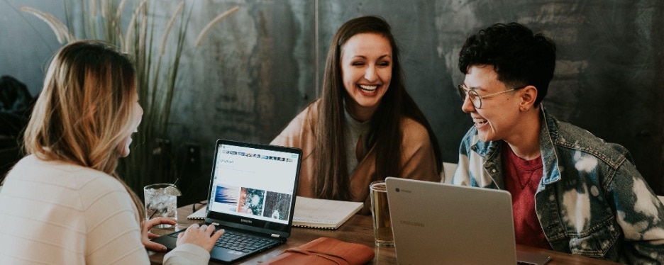Three people sitting on a picnic bench style table with laptops in front of them. They are talking and laughing with one another.