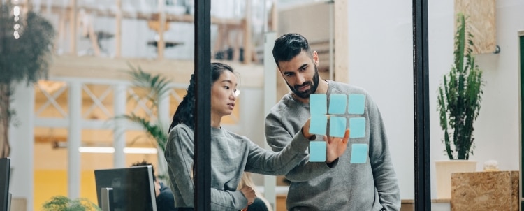 male and female coworkers wearing grey sweatshirts and pointing to blue sticky notes stuck to a clear door