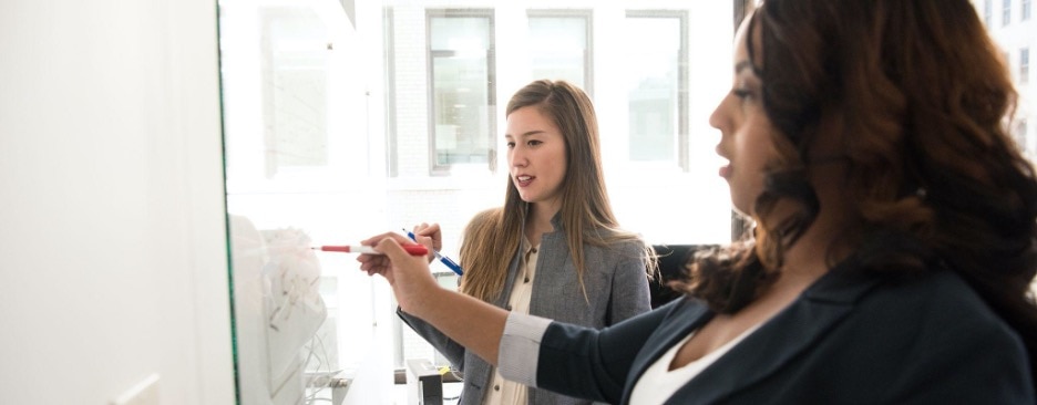 Two women in blazers writing at a whiteboard.